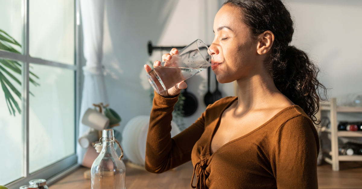 Young woman in a brown top drinking a glass of water from a carafe in her kitchen in the morning