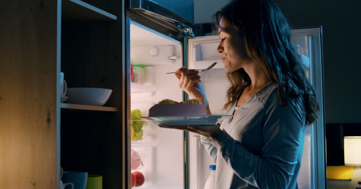 Woman smiling while enjoying cake during a midnight snack, illuminated by refrigerator light in cozy kitchen