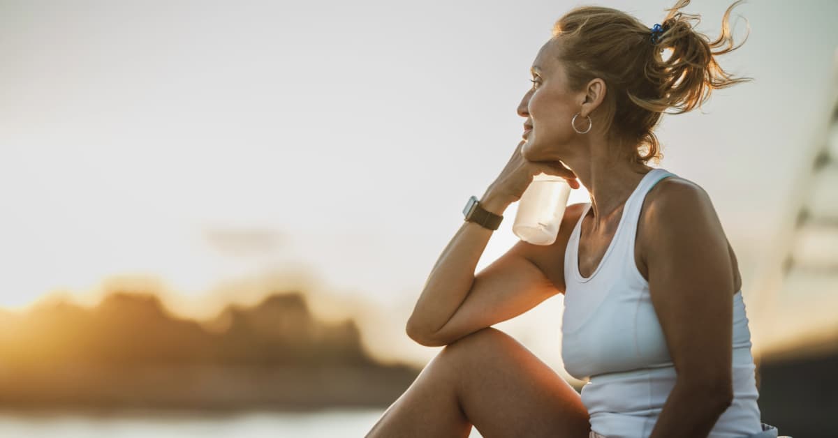 middle aged woman in white workout top holding a water bottle watching the sunset