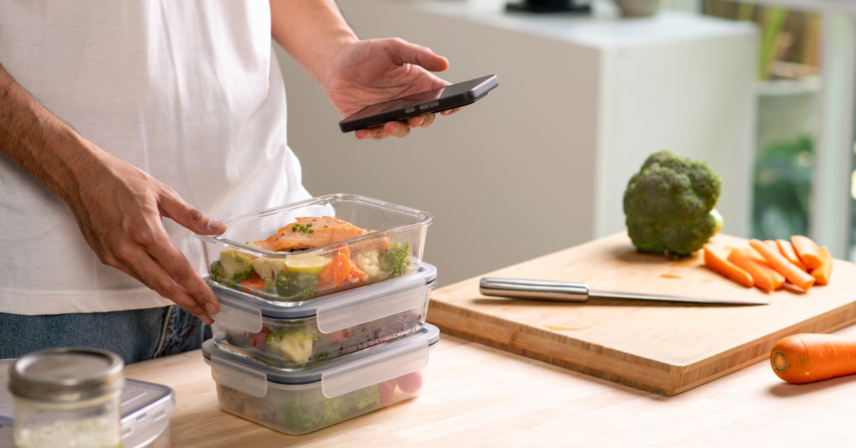 man in white shirt holding phone to track calories in meal preps sitting on his kitchen counter
